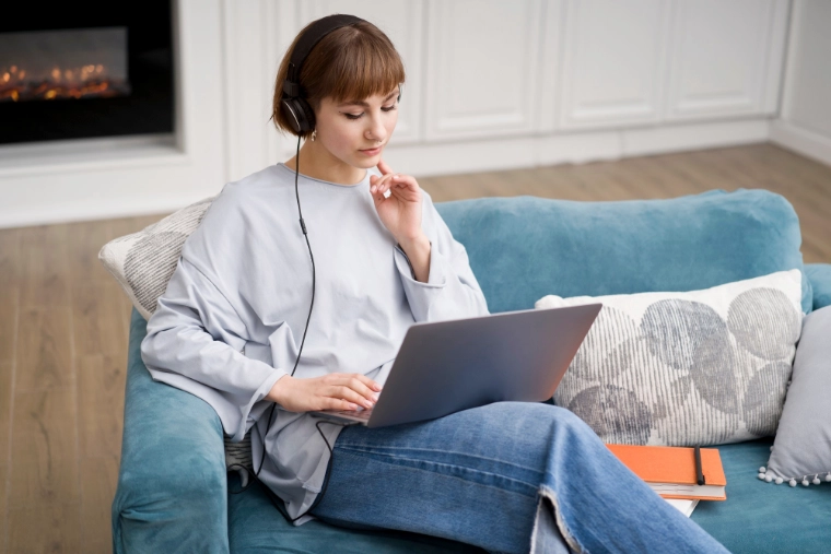 Eine Frau mit Kopfhörern sitzt entspannt auf einen blauen Sofa und schaut in einen Laptop auf ihren Knien